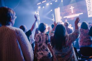 Menschen, von hinten fotografiert, Hände in der Luft. Im Hintergrund ist eine Bühne mit Kreuz und einem Banner mit der Aufschrift "Kongress" zu erkennen. Foto: spirit-kongress.de