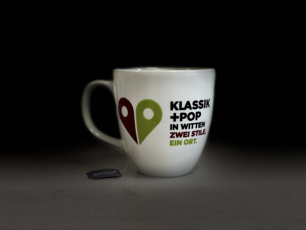 Eine Tasse mit der Aufschrift "Klassik+Pop in Witten - Zwei Stile, ein Ort". Daneben ein Herz, bestehend aus zwei Ortsmarkern wie sie in Navigationssystemen üblich sind (Foto: Hans-Martin Julius)