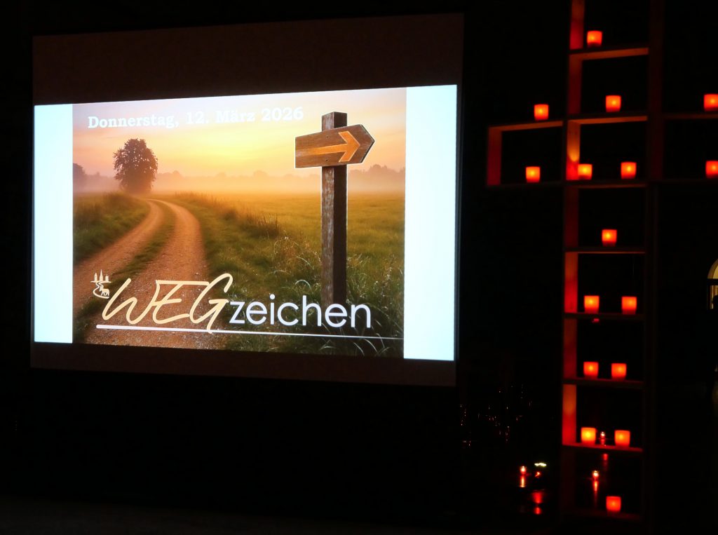 "Wegzeichen" lautet die Aufschrift auf einem Foto, das eine Morgenstimmung auf dem Land zeigt. Ein Weg führt durch eine große Wiese, aus der im ersten Licht des Tages der Nebel steigt. An einem Pfosten weist ein Pfeil den Weg. Rechts neben der Leinwand sieht man ein Kreuz aus Holzrahmen, in dem kleine Kerzen leuchten. (Foto gesamt: Hans-Martin Julius)