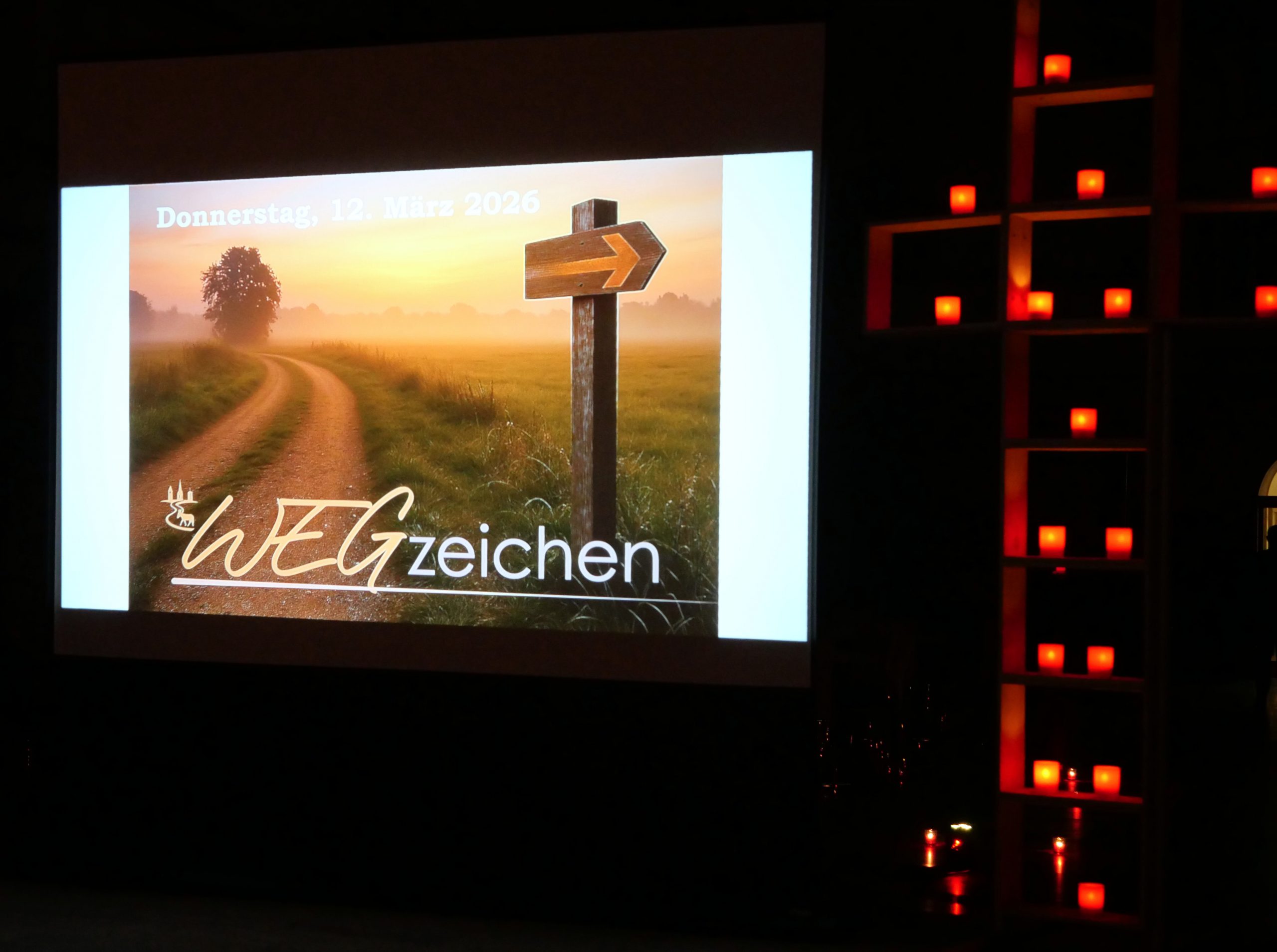 "Wegzeichen" lautet die Aufschrift auf einem Foto, das eine Morgenstimmung auf dem Land zeigt. Ein Weg führt durch eine große Wiese, aus der im ersten Licht des Tages der Nebel steigt. An einem Pfosten weist ein Pfeil den Weg. Rechts neben der Leinwand sieht man ein Kreuz aus Holzrahmen, in dem kleine Kerzen leuchten. (Foto gesamt: Hans-Martin Julius; Foto im Foto mithilfe von KI)