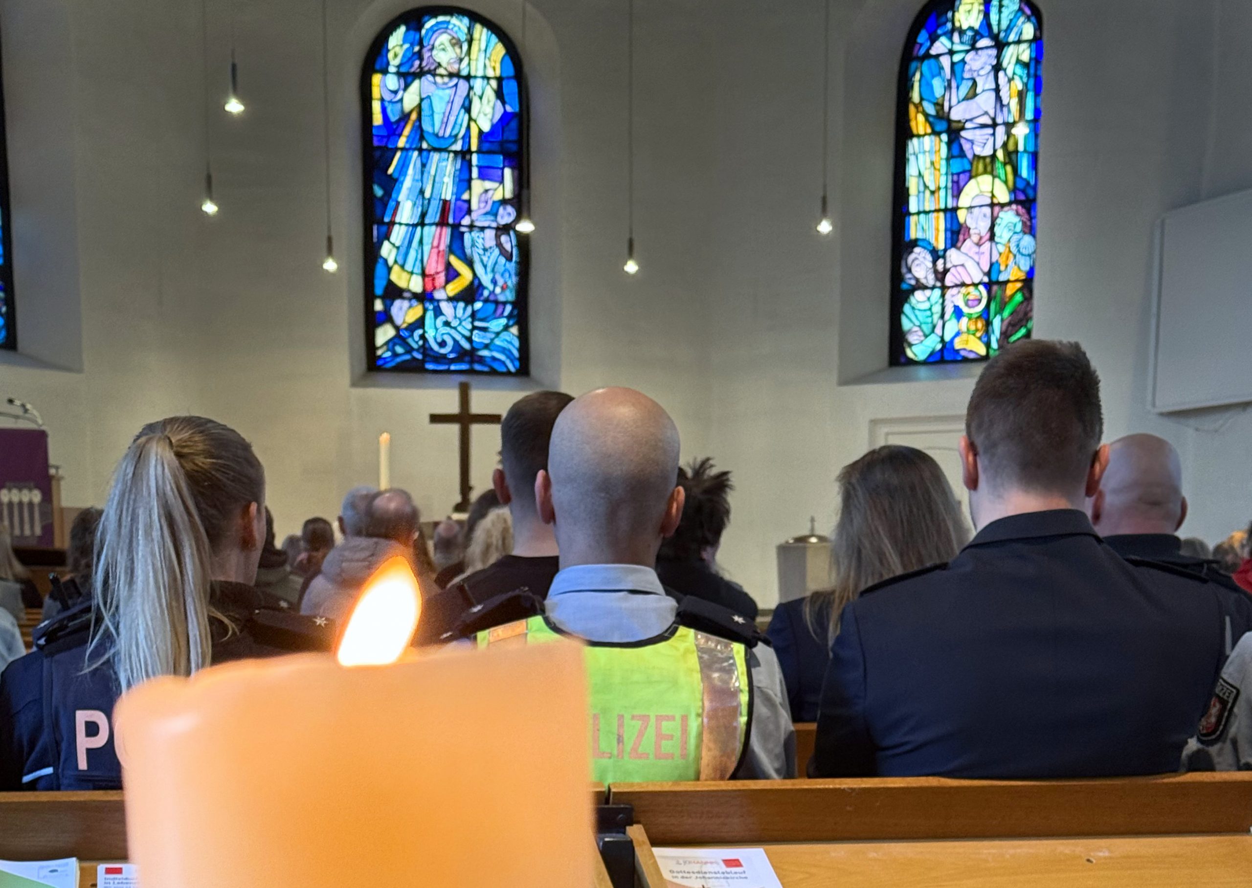 Johanniskirche Witten. Einsatzkräfte, erkennbar an ihren Westen, sitzen gemeinsam mit anderen Menschen in den Bänken der Kirche. Im Vordergrund sieht man eine Kerze, im Hintergrund Altarkreuz und Kirchenfenster (Foto: Hans-Martin Julius)